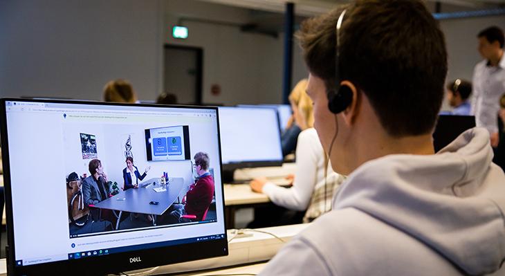 Junge Person sitzt mit Kopfhörern vor einem Monitor, auf dem ein Bild eines Verkaufsgesprächs zu sehen ist