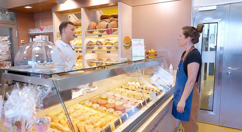 Ein Prüfling steht hinter der Theke einer Bäckerei und spricht mit einer Prüferin auf der anderen Seite der Theke