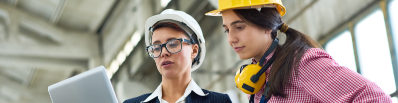 Zwei Frauen in Arbeitskleidung stehen in Fabrikumgebung und blicken auf Laptop-Bildschirm.