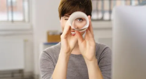 Junge Frau in Büro sitzend blickt durch gerolltes Blatt Papier